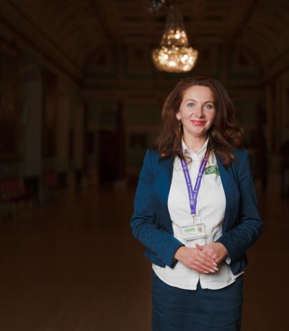 A woman with long curly hair stands in a dimly lit, ornate hall, wearing a blue blazer, white shirt, and lanyard. The hall features chandeliers and portraits on the walls.