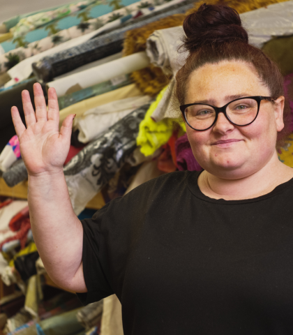 Terri wearing glasses and a black tshirt waving and smiling at the camera with colourful rolls of fabric in the background