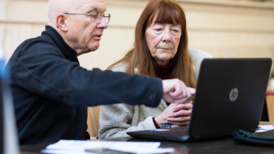 An older man and older woman sat in front of a laptop with paper on the table discussing plans.