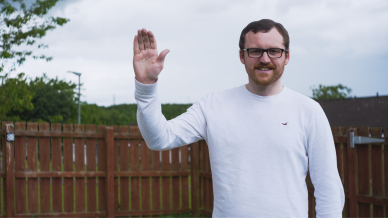Carl waving and smiling at the camera in a white long sleeved shirt while stood outside 