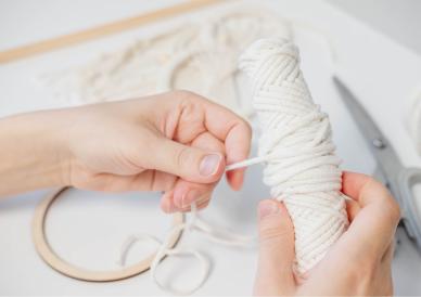 Two hands hold and wrap white string around a spool. Scissors and other crafting materials are visible in the background.