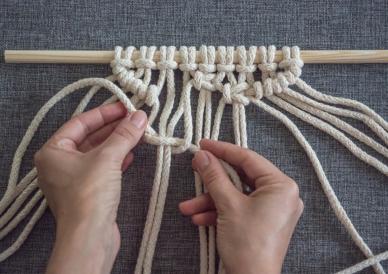 Hands are creating macramé patterns using white rope hung on a wooden dowel.