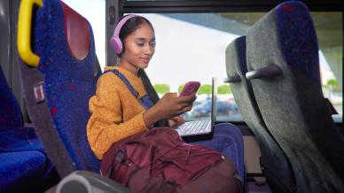Woman on the bus wearing pink headphones and holding a pink phone. She has a dark pink backpack next to her and a laptop on her lap. She is wearing a yellow jumper and denim dungareers